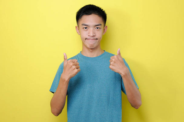 Fiatal ázsiai férfi becsukja a száját, így két hüvelykujját felfelé, elszigetelt sárga háttér - Fotó, kép