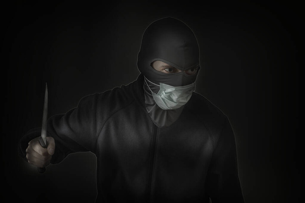Bandita v černé masce se štěrbinou pro oči a lékařskou maskou mávající nožem. Nízký průlez. Detailní záběr. Kopírovat prostor - Fotografie, Obrázek