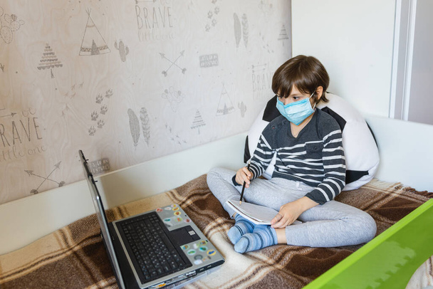 Uczenie się na odległość dla dzieci podczas epidemii koronawirusów. Chłopak siedzi na łóżku i otrzymuje zadanie szkolne za pomocą Internetu i laptopa.. - Zdjęcie, obraz