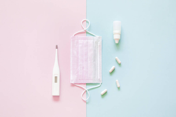 Top view of medical disposable mask, pills and a thermometer on pink and green flatlay background. Coronavirus disease or corona virus concept. Space for text - Zdjęcie, obraz