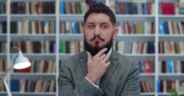 Young Caucasian businessman touching his beard and thinking over something very carefully at books shelves behind. Portrait of serious man considering some philosophy theory in library. Study concept. - 映像、動画