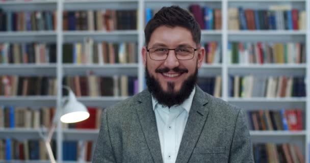 Portrait of handsome guy standing in libary and laughing cheerfully at camera. Close up of happy joyful face of male in bibliotheca. Young Caucasian man smiling indoor with books shelves on background - Filmati, video
