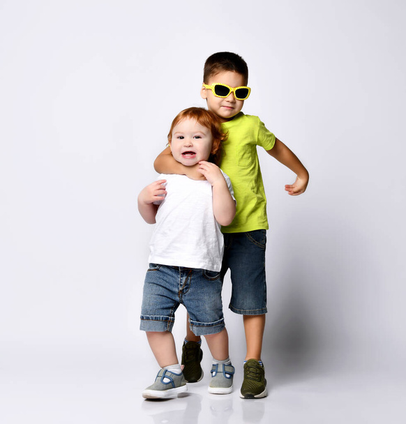 Brunet kid in sunglasses is hugging by neck his little baby brother. Smiling, dressed in casual clothes, posing isolated on white - Fotó, kép
