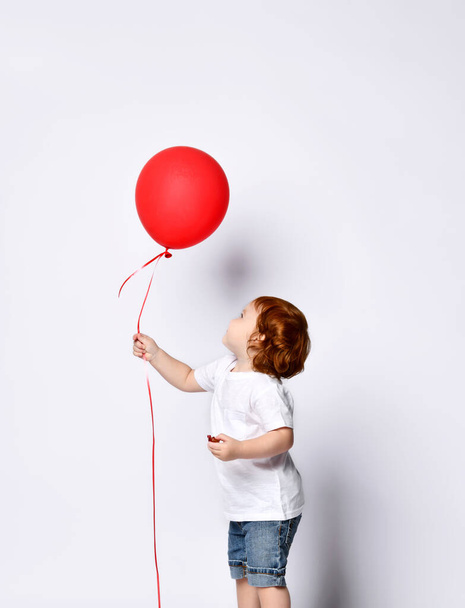 Toddler boy or girl in white t-shirt, shoes, denim shorts. Child smiling, holding red balloon, posing sideways isolated on white - Фото, изображение