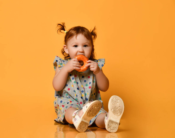 Toddler girl with two ponytails, in colorful dress and white sandals. Sitting on floor, eating donut, posing on orange background - Фото, зображення