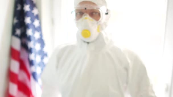 Man is holding a picture with the inscription stay at home. American doctor in a protective suit and mask on the background of the US state flag. Coronovirus covid19 and quarantine in America - Imágenes, Vídeo