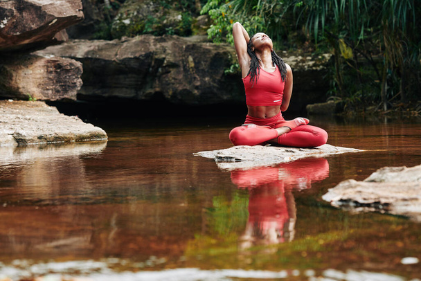 Pěkně fit mladá žena dělá jednu ruku nahoru, jednu ruku za jóga sponu, když sedí na skále v lese řeky - Fotografie, Obrázek