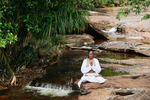 Koncentrált nő fehér laza pamut ruhát meditál lótuszpozícióban félreeső helyen a kis erdő folyó - Fotó, kép