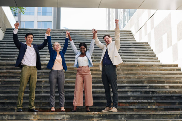 Ενθουσιασμένοι νέοι επιχειρηματίες που κρατιούνται χέρι-χέρι και σηκώνουν τα χέρια όταν στέκονται σε σκαλοπάτια έξω - Φωτογραφία, εικόνα
