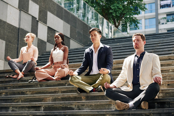 Jonge rustige zakenmannen en zakenvrouwen zittend op stappen in lotuspositie en mediterend met hun ogen dicht - Foto, afbeelding