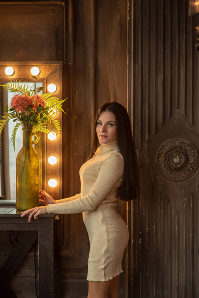 beautiful elegant brunette is standing in front of a tall make-up mirror. A girl is standing at the cosmetic table.Portrait of a young girl in a white knitted dress. - Fotó, kép