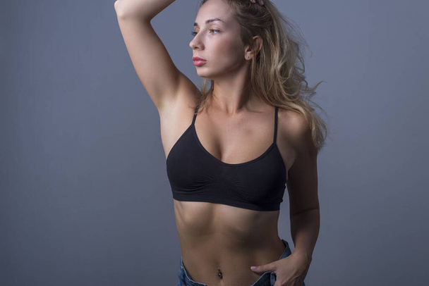 studio portrait of a young beautiful blonde girl, twenty-eight years old. In jeans and a black bra top. Advertising photo on a gray background. Free space. - Foto, Imagen