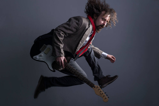 defocused studio portrait of a young bearded, curly-haired guy with long hair, in a jacket, jumping with a guitar, looking at the camera. On a gray background. Dynamic male dramatic portrait - Photo, Image