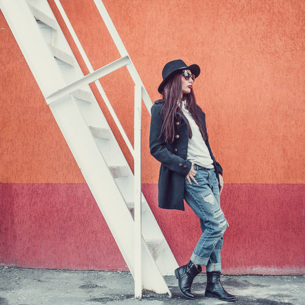 young beautiful girl with bright lips, stylishly dressed, on her head a black hat with brim and glasses, she is wearing a black coat and jeans, she is standing against a wall next to a steel staircase - Photo, image