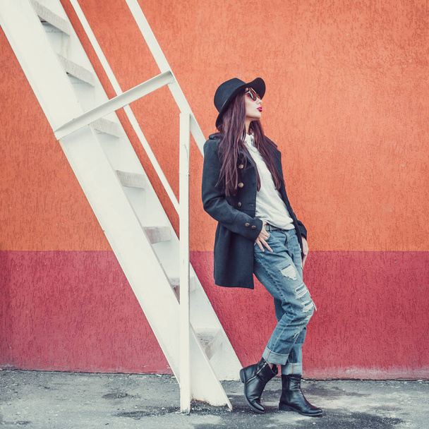 young beautiful girl with bright lips, stylishly dressed, on her head a black hat with brim and glasses, she is wearing a black coat and jeans, she is standing against a wall next to a steel staircase - Foto, Imagem