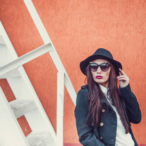 young beautiful girl with bright lips, stylishly dressed, on her head a black hat with brim and glasses, she is wearing a black coat and jeans, she is standing against a wall next to a steel staircase - Foto, Imagen
