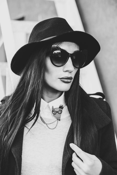 young beautiful girl with bright lips, stylishly dressed, on her head a black hat with brim and glasses, she is wearing a black coat and jeans, she is standing against a wall next to a steel staircase - Foto, imagen