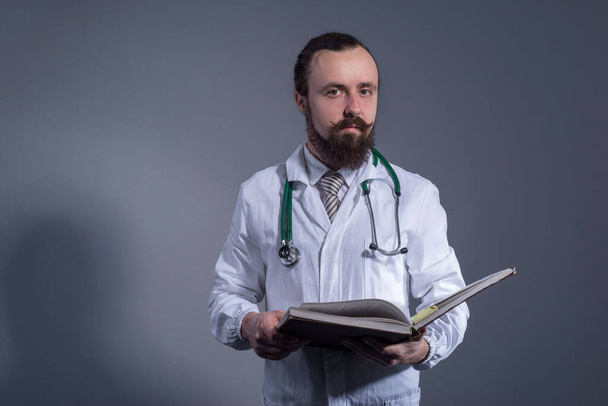 Προσωπογραφία γενειοφόρου γιατρού με λευκό παλτό και φωνοενδοσκόπιο που κρατάει στα χέρια του εκπαιδευτικά βιβλία. Φωτογραφία στούντιο σε γκρι φόντο. Αυτο-εκπαίδευση του γιατρού. Ιατρική γνώση, η δύναμη της γνώσης. Επαγγελματική επιμόρφωση των ιατρών - Φωτογραφία, εικόνα