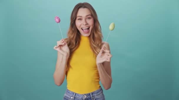 Cheerful young girl shakes easter eggs isolated on blue background - Metraje, vídeo