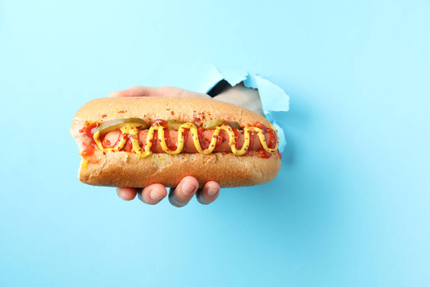 Hand holds hot dog from hole on blue background - Foto, imagen