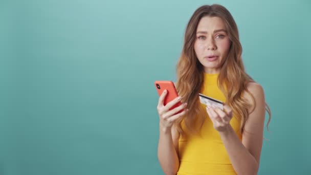 Sad young girl shopping online and paying for purchases using credit card and smartphone on isolated blue background - Video