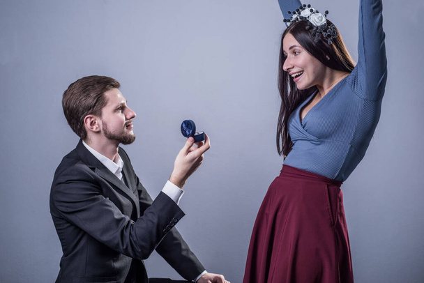Studioporträt eines Kerls kniend, macht einem sehr glücklichen Mädchen einen Heiratsantrag mit einem Ring. Auf grauem Grund. Heiratsantrag mit Herz und Hand, heiraten, heiraten. Verlobung, Verlobungsring. Konzept - Foto, Bild