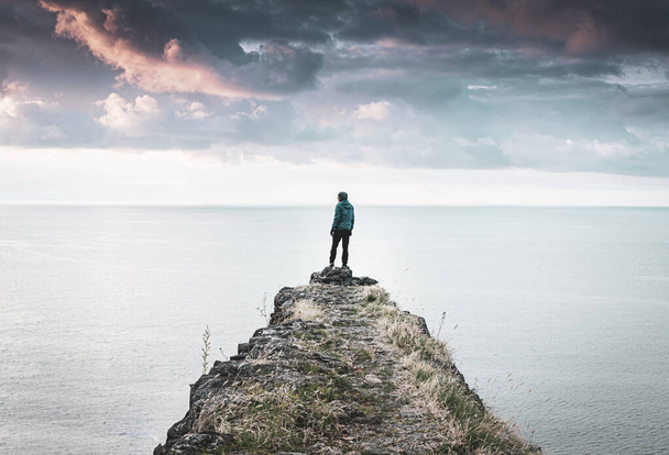 Person stands alone and looking around on the edge of  the cliff with vast sea and dramatic sky in the background.  Concept of solitude, adventure and lifestyle. 2020 - Fotoğraf, Görsel