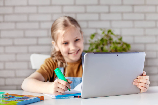 Een vrolijk meisje voert huiswerk met een boek en een tablet thuis. Training thuis, zelfisolatie, quarantaine thuis. Technologie, onderwijs, online leren, afstandsonderwijs thuis - Foto, afbeelding