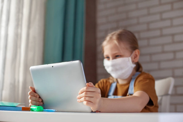 Una chica con una máscara médica está estudiando en casa con un portátil tableta digital y haciendo la tarea. Educación a distancia en línea
 - Foto, Imagen