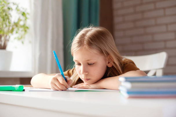 Egy gyönyörű lány tollal a füzetében ír. A gyerek házit csinál. Otthoni képzés, online képzés - Fotó, kép