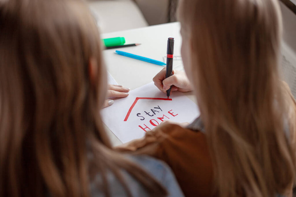 Οι αδερφές ζωγραφίζουν μια ταμπέλα στο χαρτί για να μείνουν σπίτι. Πρόληψη, κοινωνική απόσταση, ηλεκτρονική μάθηση, εξ αποστάσεως εκπαίδευση - Φωτογραφία, εικόνα