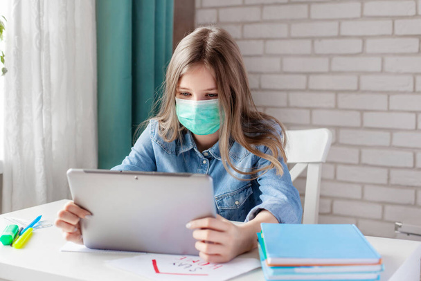医療用マスクの女の子は、デジタルタブレットのラップトップで自宅で勉強して宿題をしています。遠隔オンライン教育 - 写真・画像