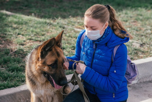A girl in a protective medical mask walks a dog on the street. Leisure with a pet during quarantine. Walk with a German shepherd in the fresh air. Self-isolation and protection mode. - Fotografie, Obrázek