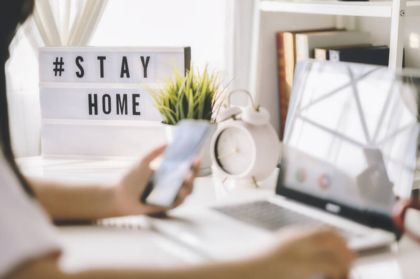 lightbox with text hashtag #STAYHOME glowing in lightand blurred woman working at home. Office worker on quarantine. Home working to avoid virus disease. Freelancer or remote worker concept.  - Photo, Image