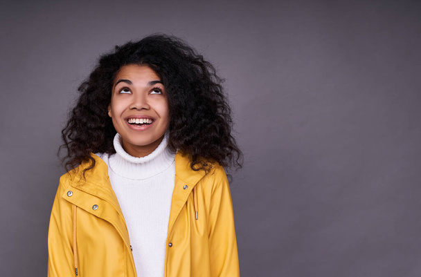Photo of a mixed race, a girl with magnificent curly hair, in a yellow raincoat sends a kiss. Studio shot of a wonderful curly woman on a gray background. Copy space. - Photo, Image