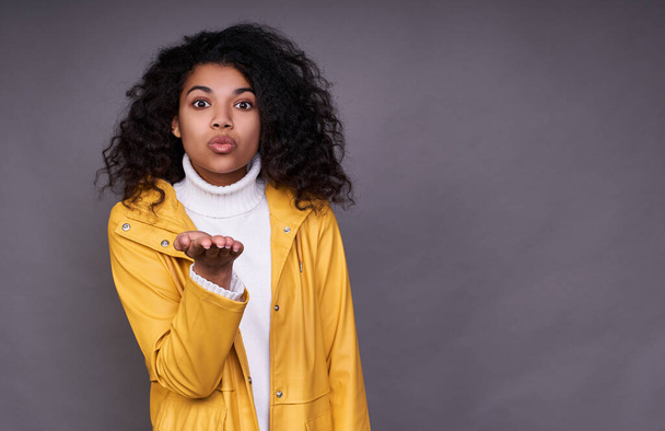 Photo of a mixed race, a girl with magnificent curly hair, in a yellow raincoat sends a kiss. Studio shot of a wonderful curly woman on a gray background. Copy space. - Foto, afbeelding