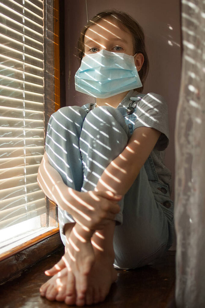 Väsynyt tylsistynyt pikkutyttö istuu ikkunalaudalla suojaava kertakäyttöinen naamio hänen kasvonsa aikana itsensä eristäminen kotona, koska Coronavirus Covid-19 kaihtimet tausta
. - Valokuva, kuva