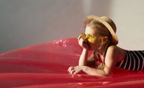 Happy little child girl in a swimsuit and a straw hat prepares for a summer trip at home.  Lies on an air mattress and dreams of quarantined summer - Foto, Imagem
