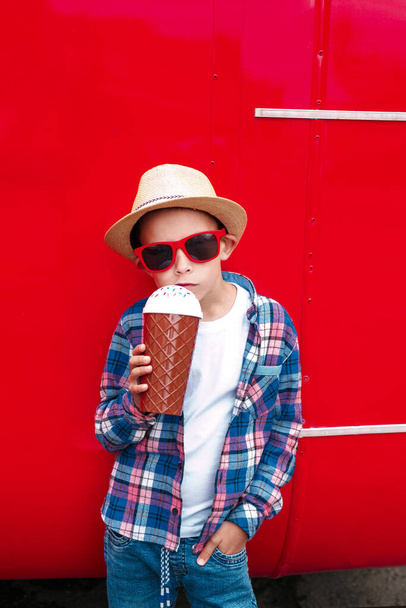 Little boy in sunglasses and hat is drinking milk cocktail over red city background - Φωτογραφία, εικόνα