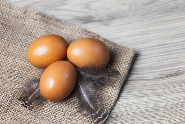 Tres huevos marrones con plumas en el saco.Huevos sobre fondo de madera
 - Foto, Imagen