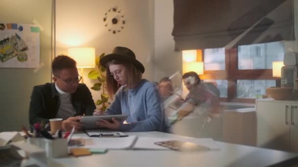 Jonge stijlvolle ontwerpers zijn het bespreken van een project op een tablet door te tekenen met een pen, achtergrond vrouw en man met laptop. - Video