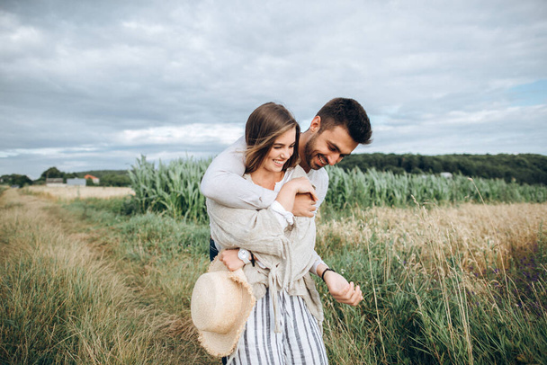 Ευτυχισμένο ζευγάρι ερωτευμένο να αγκαλιάζει, να φιλάει και να χαμογελάει στον ουρανό στο χωράφι. Καπέλο στο χέρι του κοριτσιού - Φωτογραφία, εικόνα