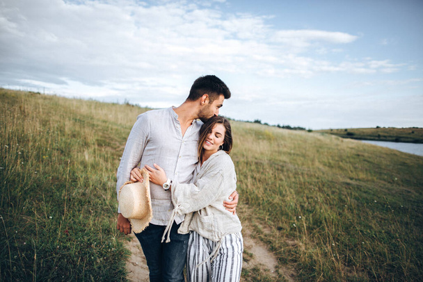 Couple heureux amoureux étreignant, embrassant et souriant contre le ciel dans les champs. Chapeau dans la main de la fille
 - Photo, image