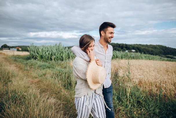 Happy couple in love hugging, kissing and smiling against the sky in field. Hat in girl's hand - Photo, Image
