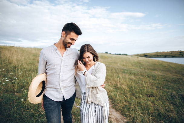 Couple heureux amoureux étreignant, embrassant et souriant contre le ciel dans les champs. Chapeau dans la main de la fille
 - Photo, image