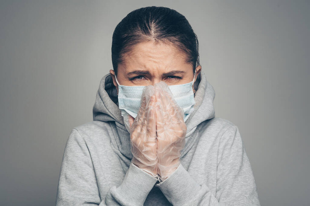 Studio portrait of young woman wearing a face mask and gloves coughs, looking at camera, on gray background. Coronavirus pandemic, dust allergy, protection against virus concept - Fotoğraf, Görsel