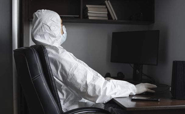 Man in medical mask and protective suit, rubber gloves sits at home and works with pc at the table during quarantine. Designer, artist, architect, businessman at remote work in a pandemic covid. - Fotoğraf, Görsel