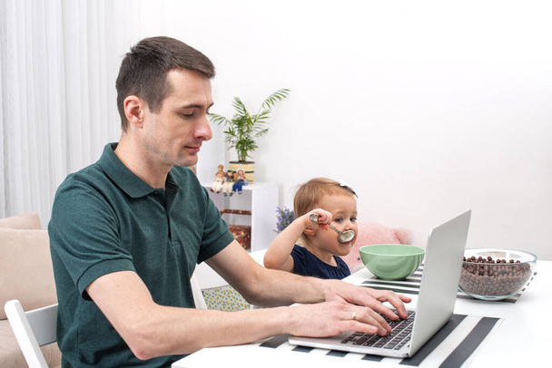 Dad take care of a little daughter at home.  He gave her cereal with milk and started working using a laptop. Life style.  The modern role of men in the family. - Photo, Image
