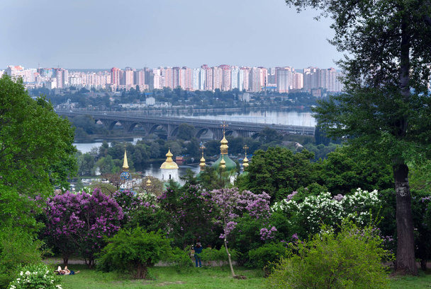 Orthodoxe kerk op de achtergrond van de rivier en de stad, in de tuin van bloeiende seringen, Kiev, Oekraïne - Foto, afbeelding
