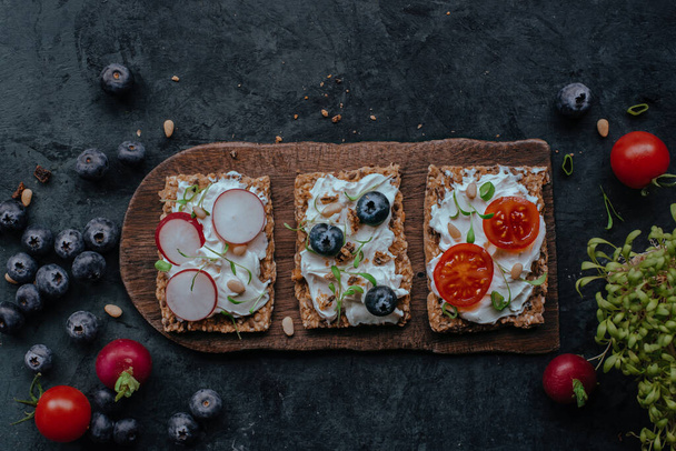 Delicioso pan crujiente multigrano con rodajas de tomate de queso crema, blubery, rábano, semillas y brotes en el tablero de madera
 - Foto, imagen